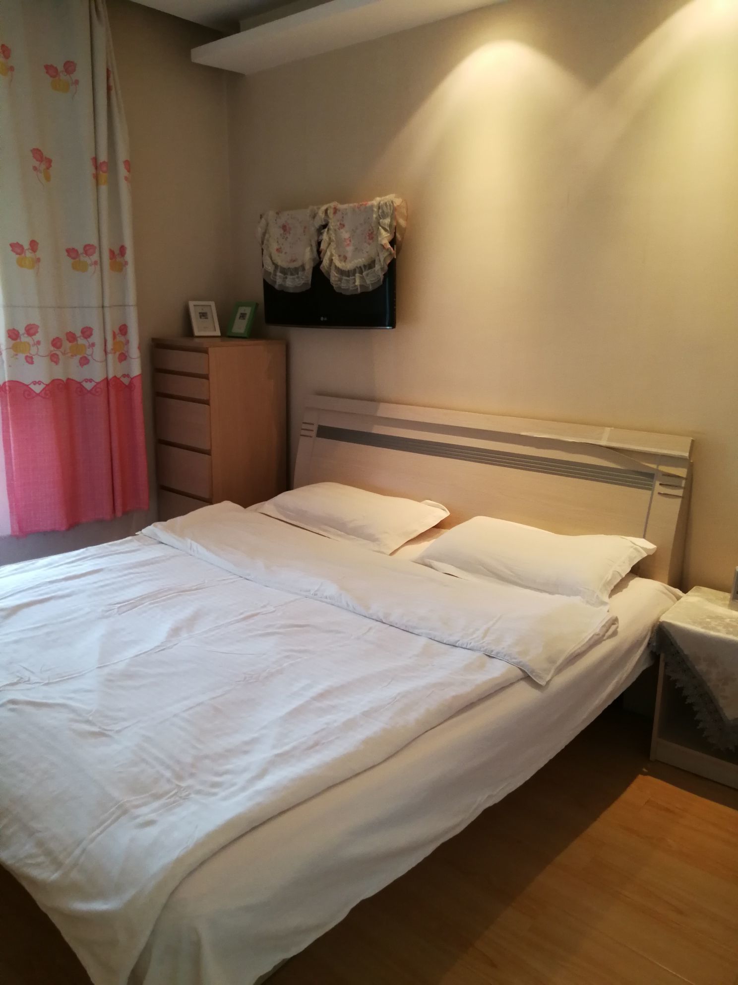 A neatly made bed with white linens, a wooden nightstand with a lamp, and a wall-mounted TV above a dresser.