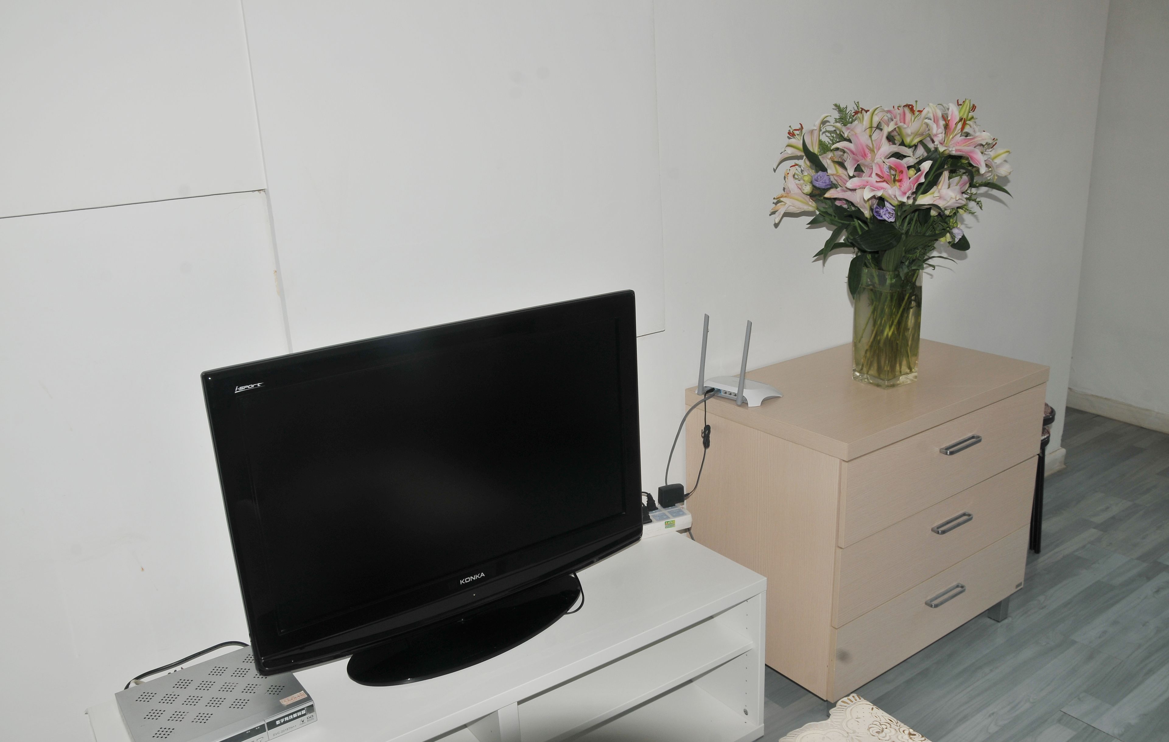 Un televisor de pantalla plana sobre un soporte blanco, un aparador beige con cajones y un florero con flores.