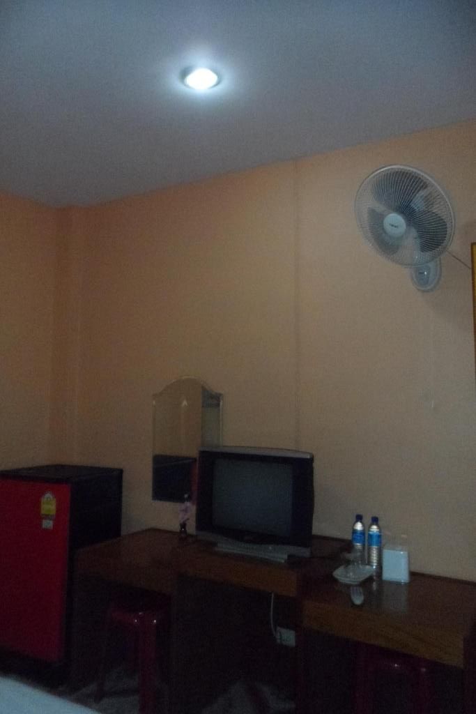 A room with a desk, a television, a refrigerator, a fan, and a mirror.
