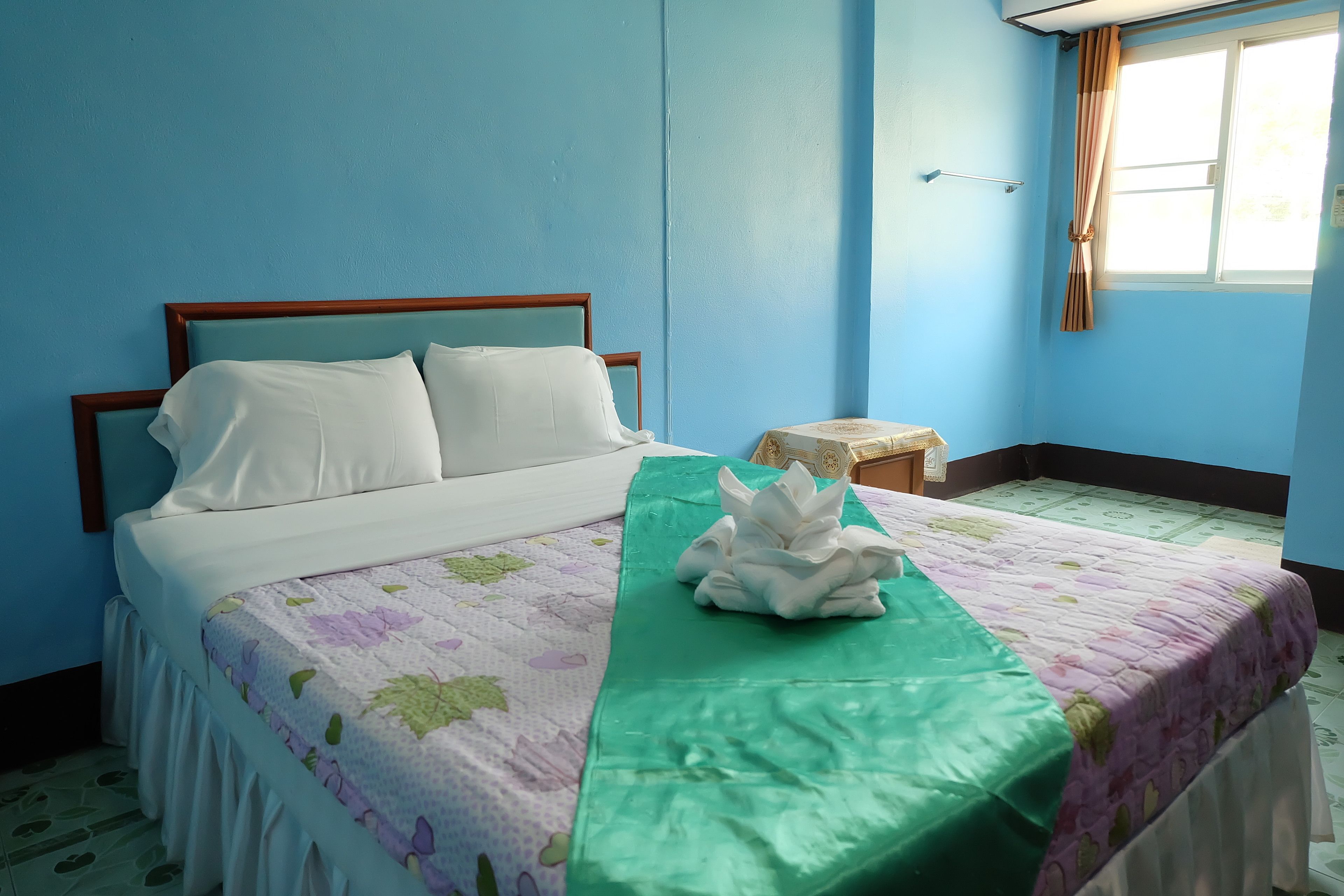 A neatly made bed with white linens and a floral-patterned bedspread. There is a green towel on the bed and a small wooden stool beside it.
