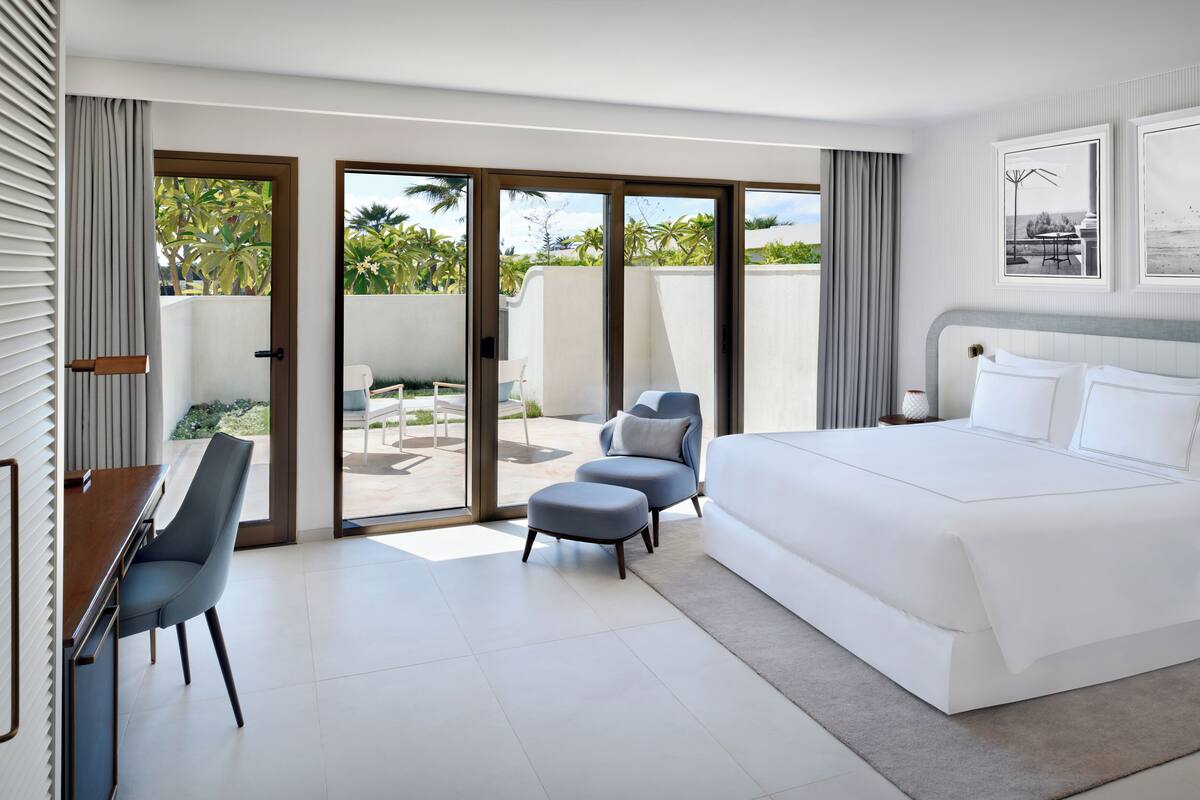 A modern hotel room with a large bed, a desk, and a chair. There is a sliding glass door leading to a balcony with a view of palm trees.