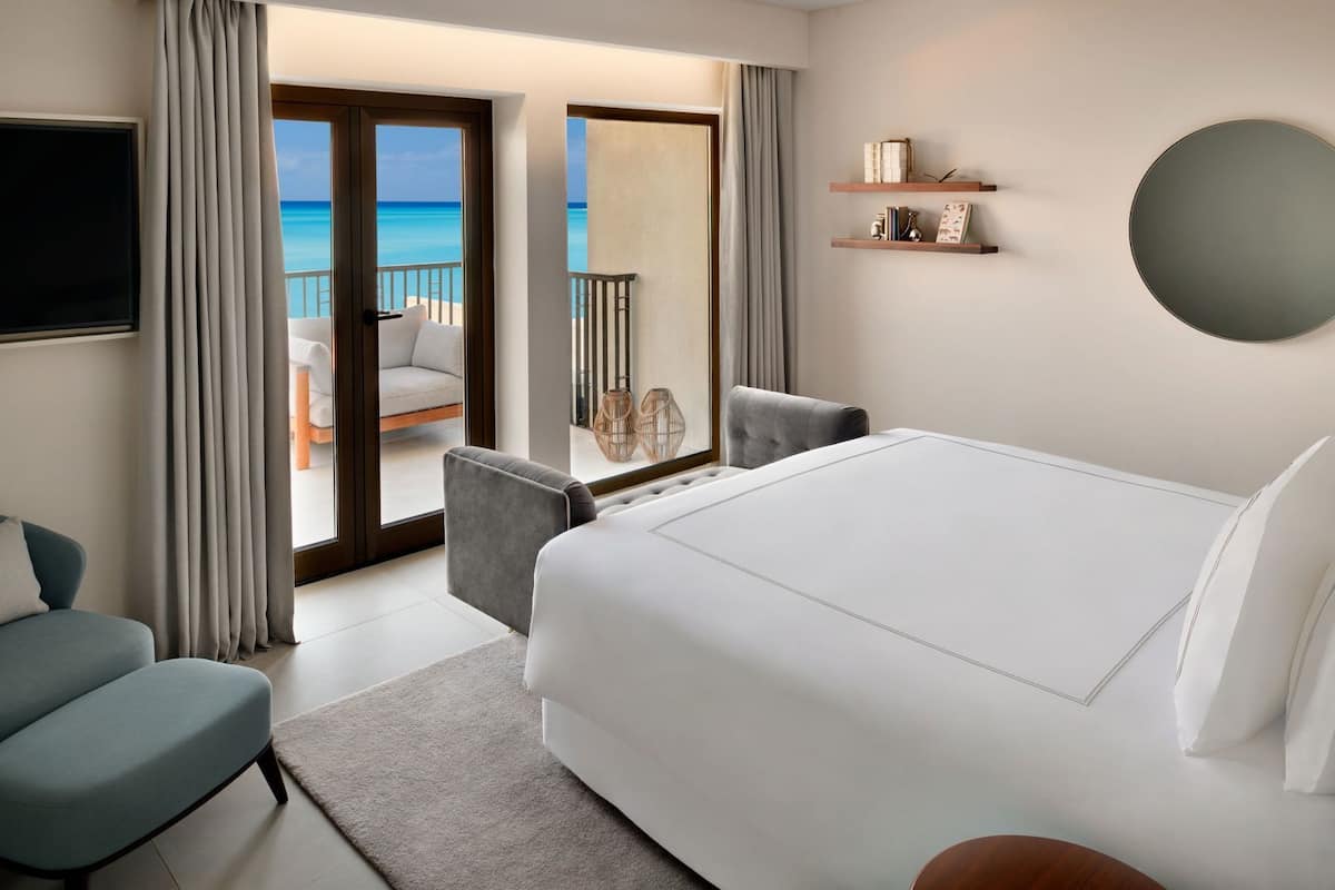 A hotel room with a large bed, a TV, a balcony with a view of the ocean, and a small seating area.