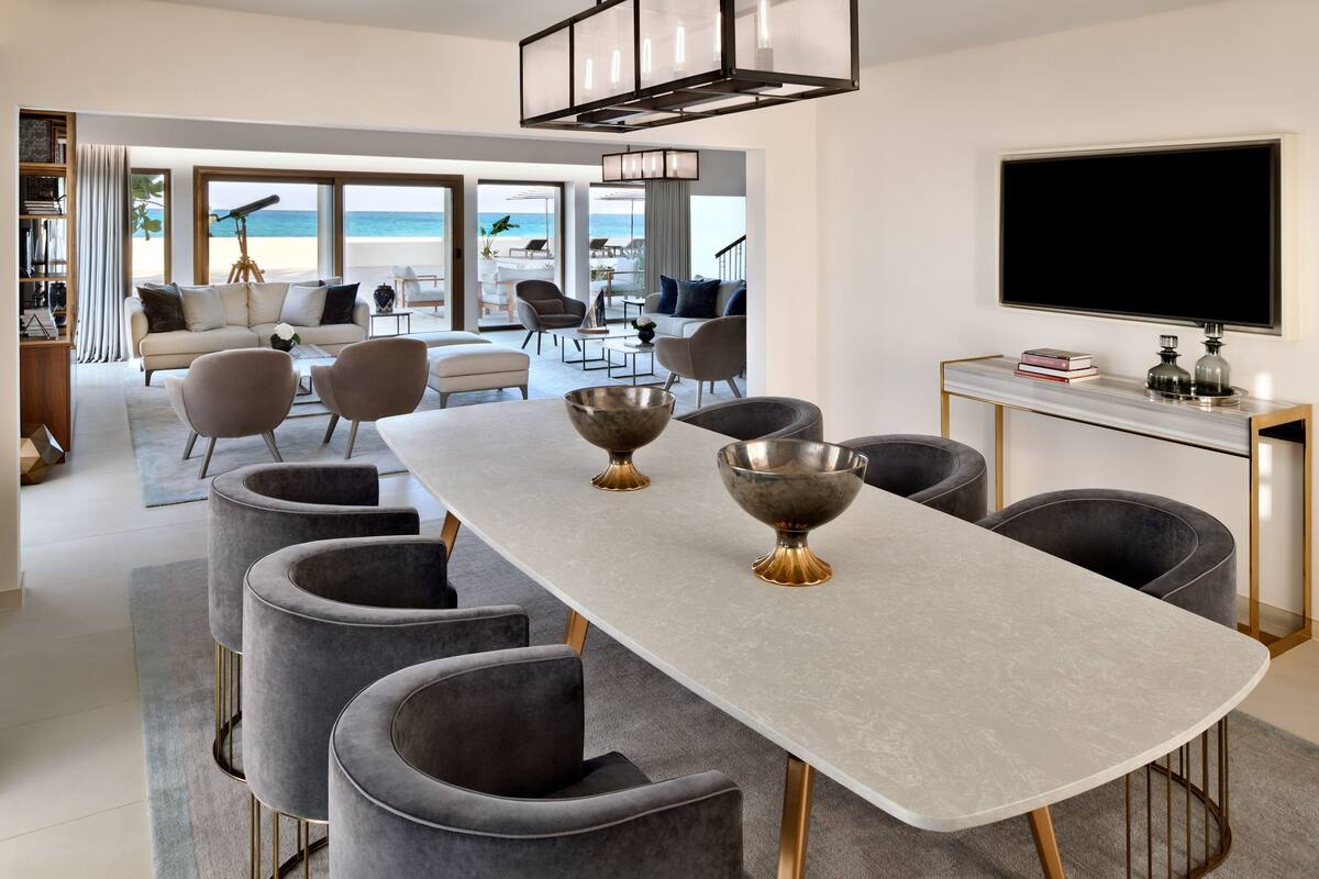 A modern living room with a long dining table, grey upholstered chairs, and a large window overlooking the beach.