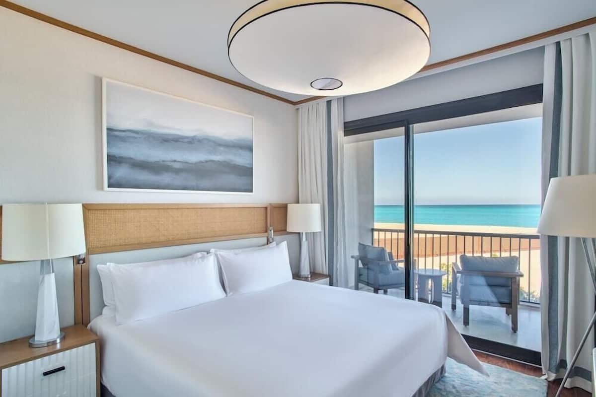 A hotel room with a bed, two bedside lamps, a large window with a view of the beach, and a ceiling light fixture.
