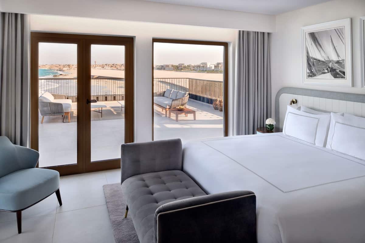 A hotel room with a large bed, a grey armchair, a blue armchair, a balcony with a view of the beach and cityscape, and a framed picture on the wall.