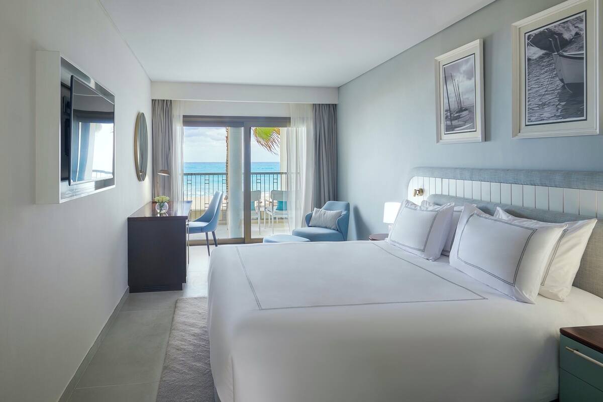 A hotel room with a large bed, a desk, a chair, and a balcony with a view of the sea.