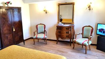 A hotel room with a wooden wardrobe, two armchairs, a small wooden dresser, a mirror, and a television.