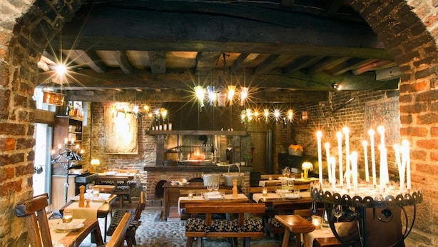 A cozy restaurant with brick walls, exposed beams, and a central fireplace. Tables are set with candles and tableware. - Auberge Du Bon Fermier (Valenciennes)