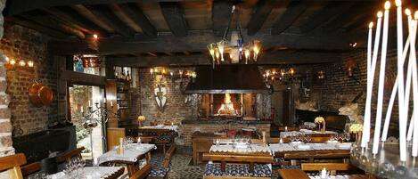 A rustic interior with exposed brick walls, wooden beams, and a central fireplace. Tables are set with wine glasses and candles, and there are benches and chairs for seating.