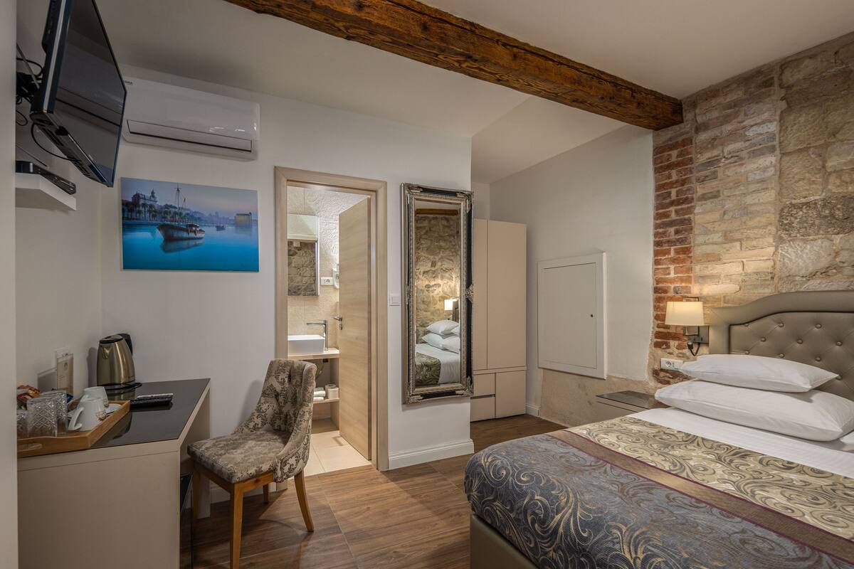 A hotel room with a bed, a chair, a desk with a kettle, a painting, a mirror, and a stone wall.