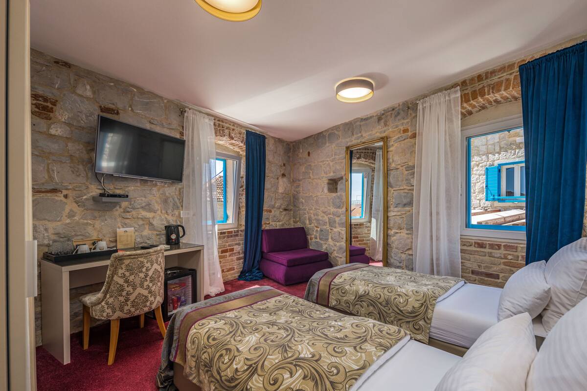 A room with two beds, a desk, a chair, a TV, and a stone wall.