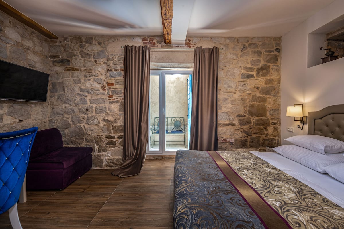 A room with a stone wall, a bed with a patterned bedspread, a blue chair, and a television on the wall.
