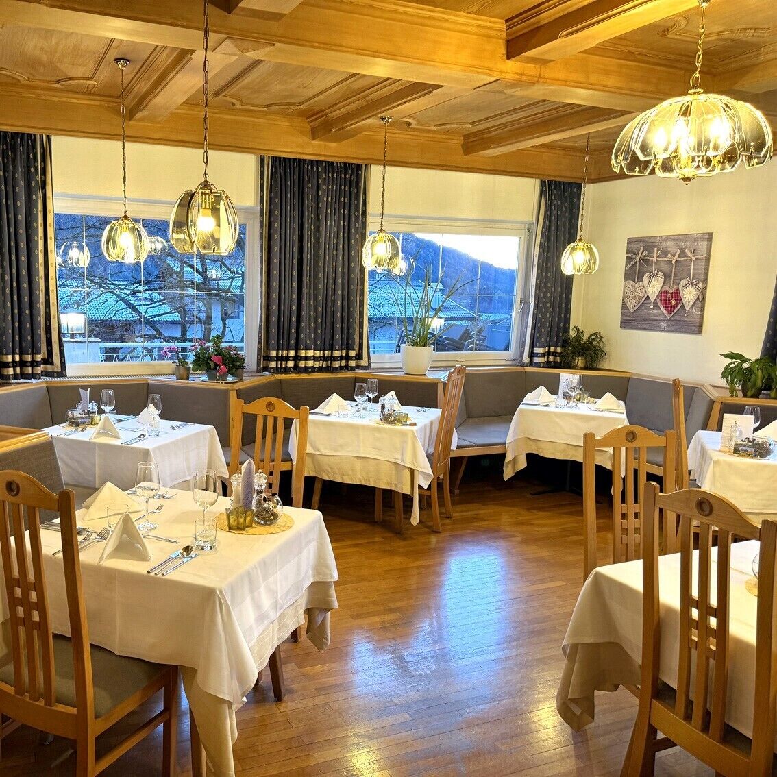 Ein Restaurant mit Holztischen und -stühlen, hängenden Pendelleuchten und großen Fenstern mit Vorhängen.