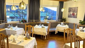Ein Restaurant mit Holztischen und -stühlen, hängenden Pendelleuchten und großen Fenstern mit Vorhängen.