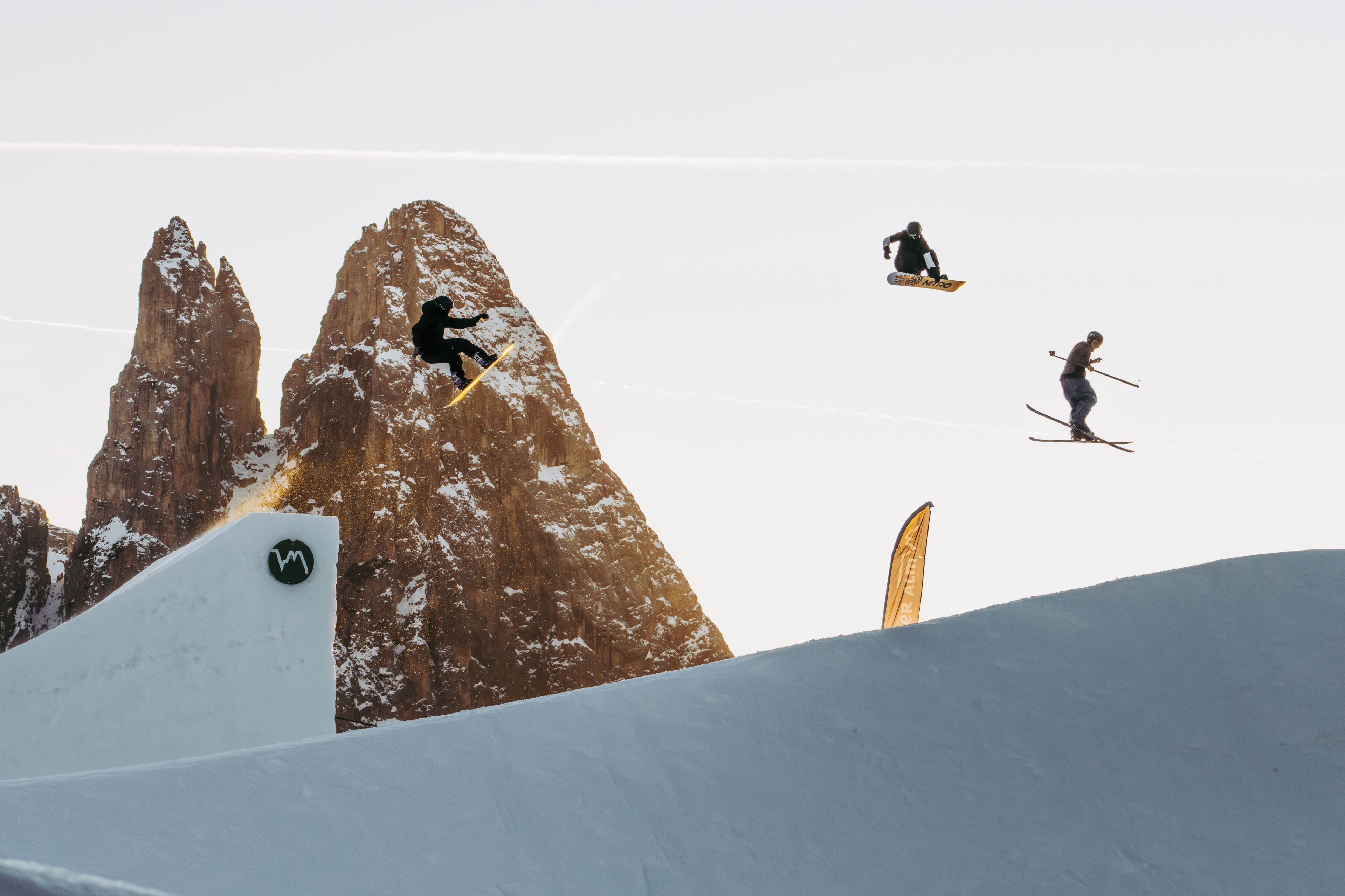 A ski slope with skiers performing jumps and a mountain backdrop.