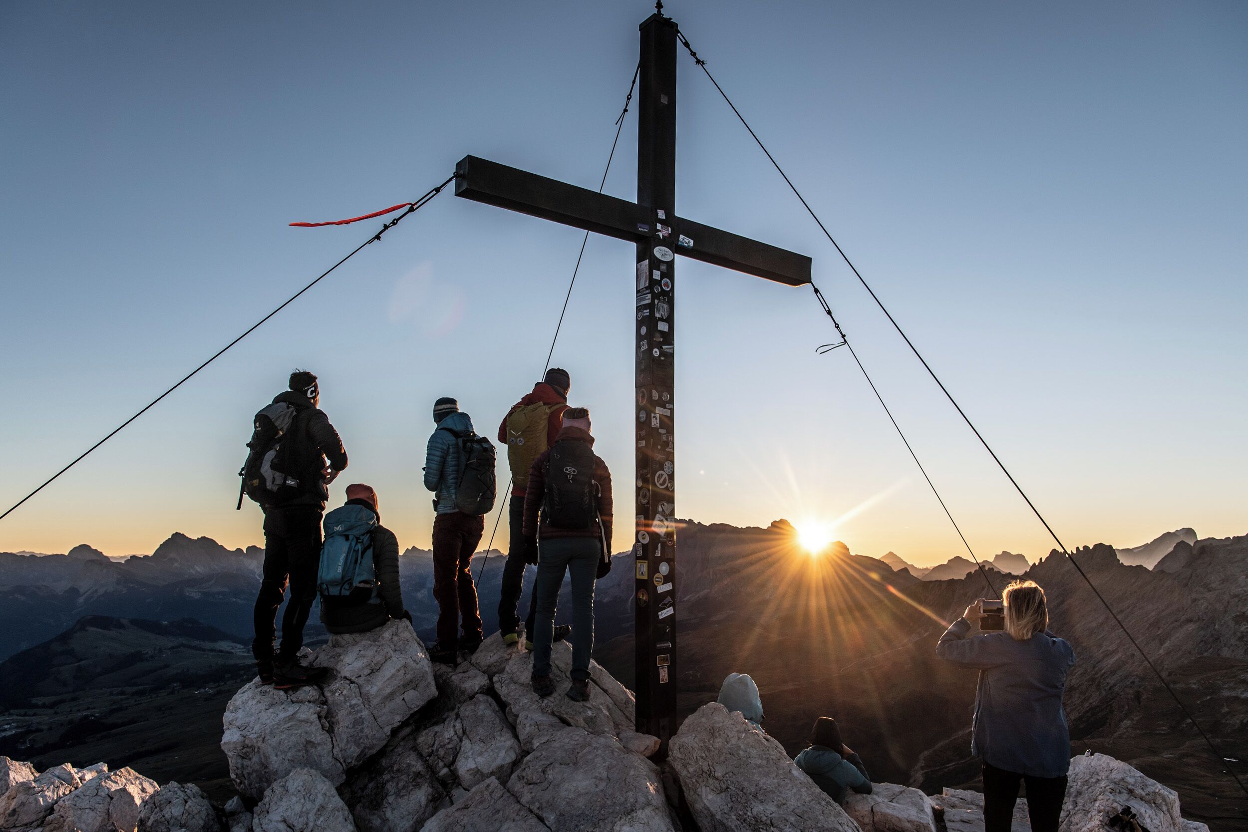 Un grup de persones al cim d'una muntanya amb una estructura de creu, en un paisatge rocosos i amb vistes al capvespre.