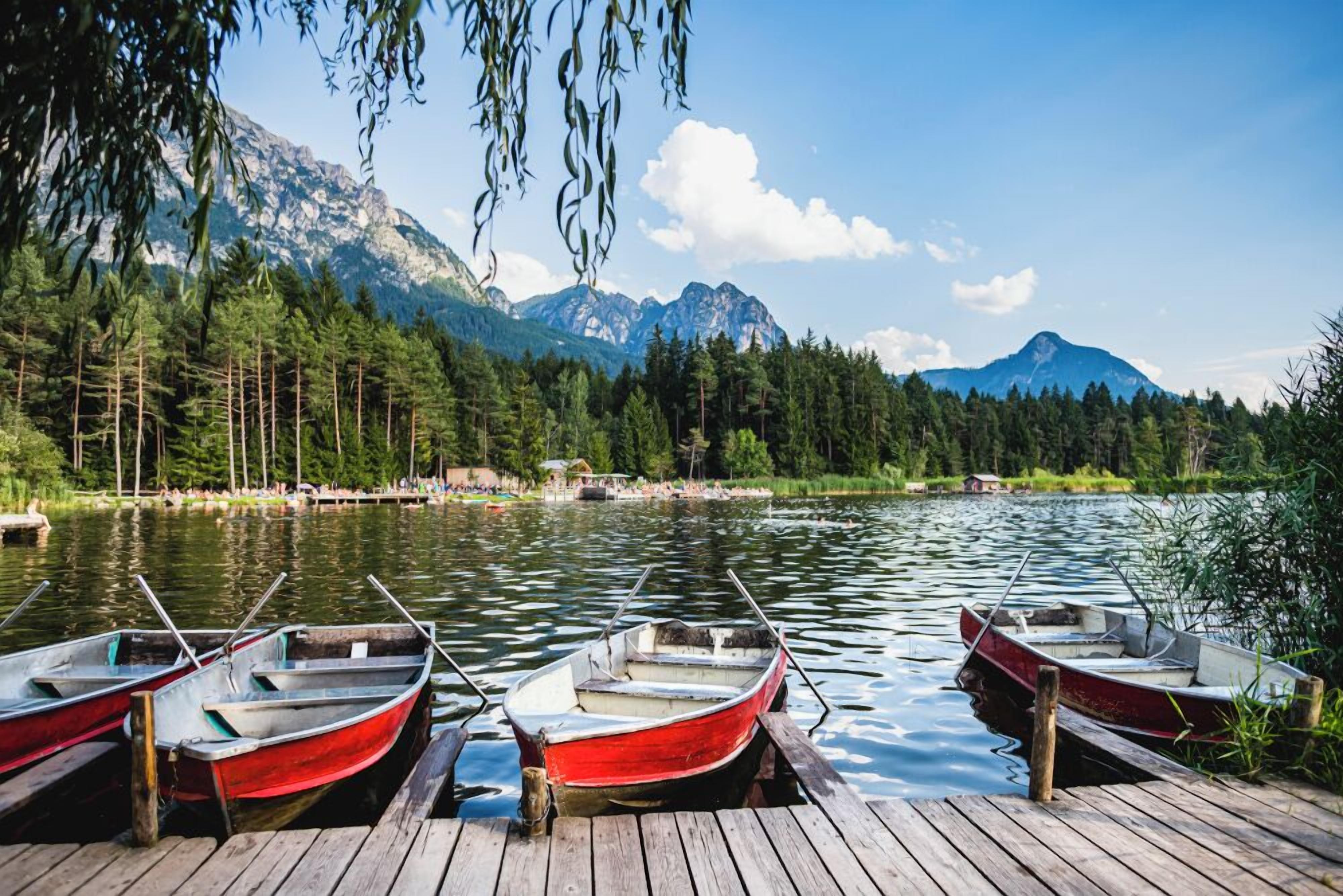 A lake with boats docked on a wooden pier, surrounded by dense forest and mountains.