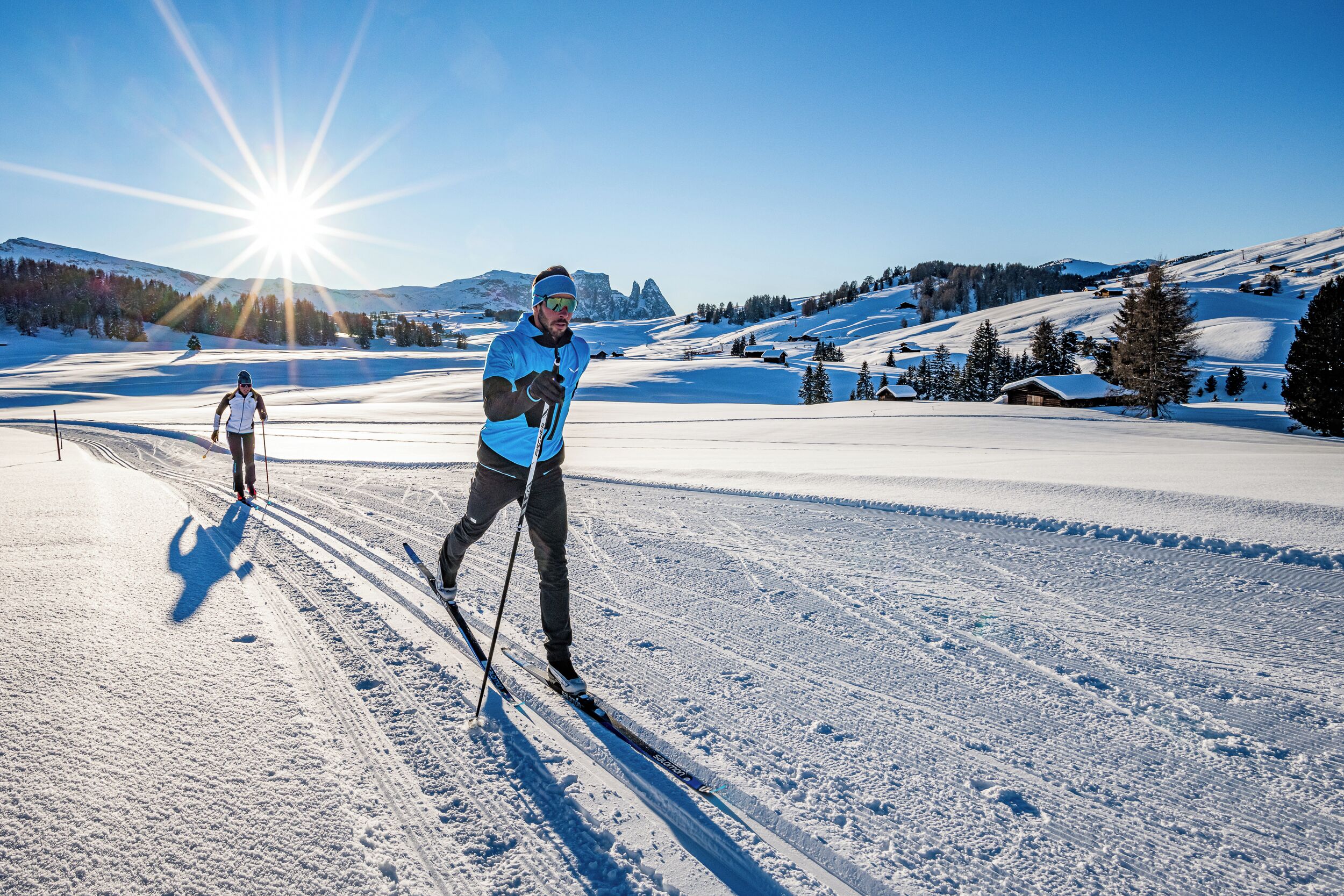 Eine Person Langlauft in einer schneebedeckten Loipe unter blauem Himmel mit Bergen im Hintergrund.