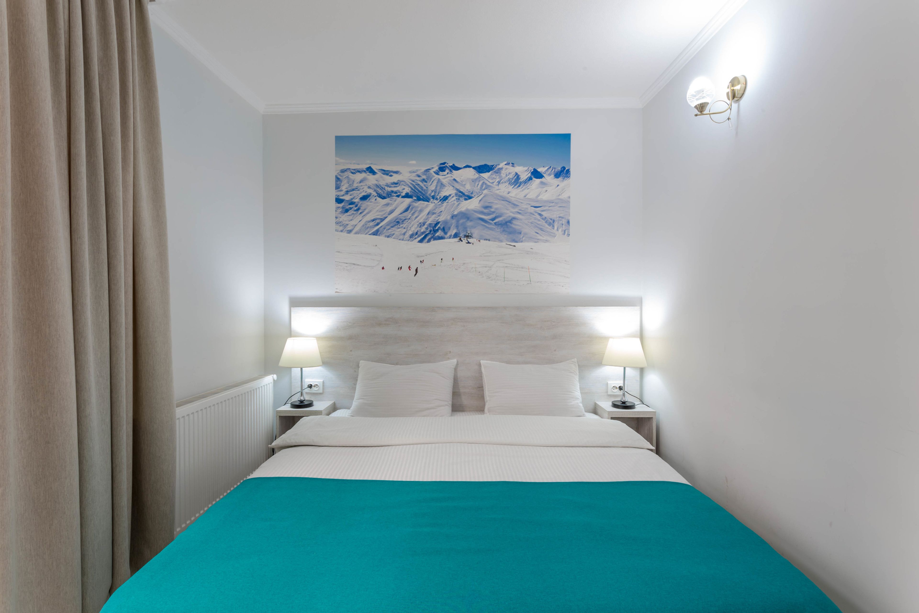 A hotel room with a bed, two bedside lamps, a headboard with a painting, and a window with curtains.