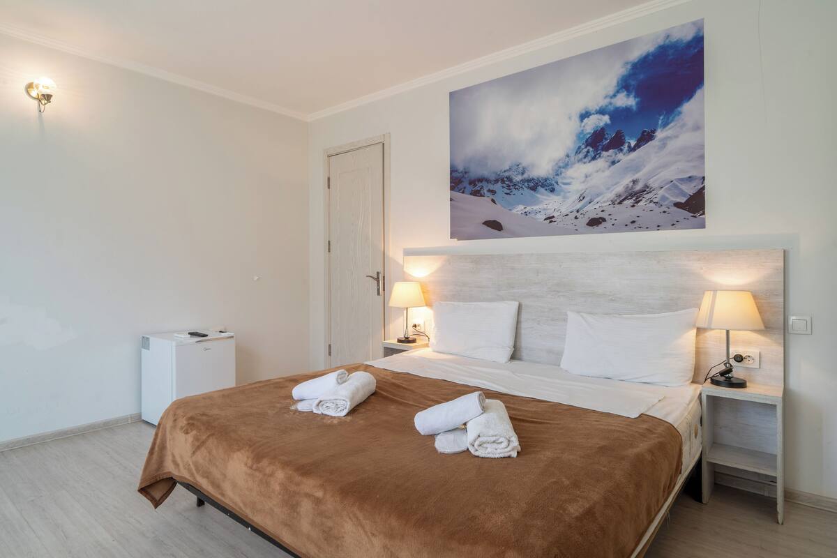 A hotel room with a bed, two bedside lamps, a nightstand, a wall-mounted picture, and a small desk with a lamp.