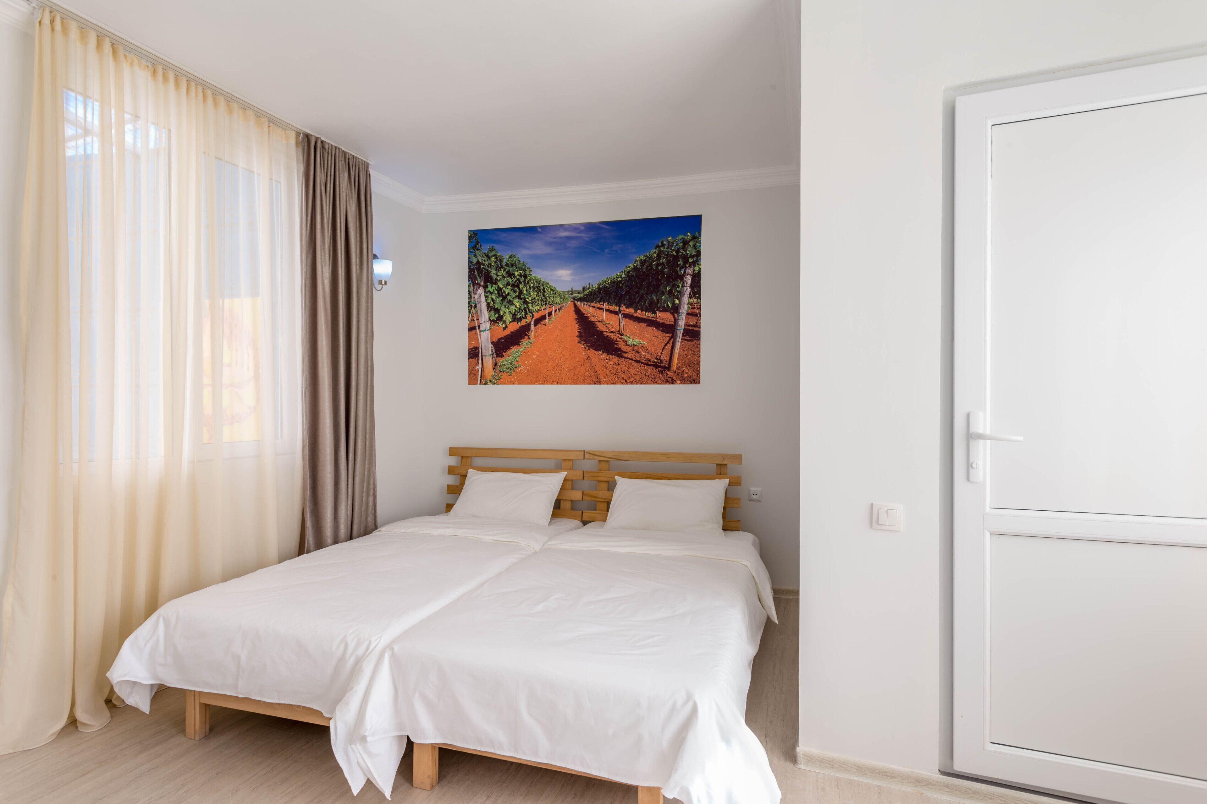 Ein Schlafzimmer mit einem Bett, einem Weinbergsgemälde, einer Tür und einem Fenster mit Vorhängen.