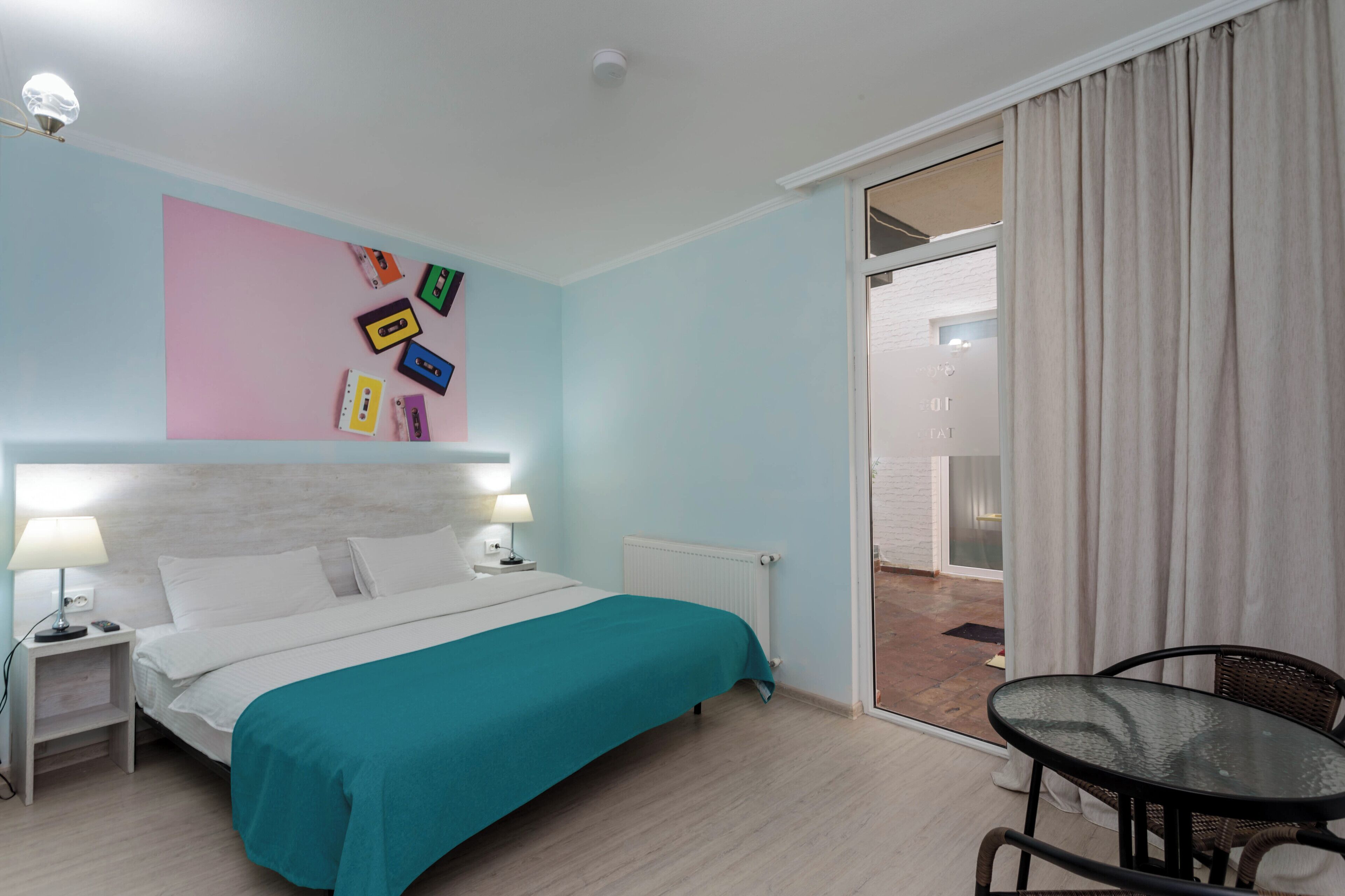 Une chambre d’hôtel avec un lit, des tables de chevet, une couvre-lit turquoise, un mur décoré de cassettes audio colorées, une porte-fenêtre coulissante donnant sur un balcon et une petite table avec des chaises.