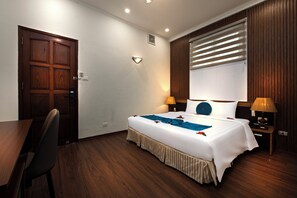 Quarto de hotel com porta de madeira, uma cama com roupa de cama branca, duas luminárias de mesa e uma janela com persianas.