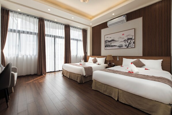 Quarto de hotel com duas camas, uma escrivaninha, uma cadeira e uma janela grande com cortinas.