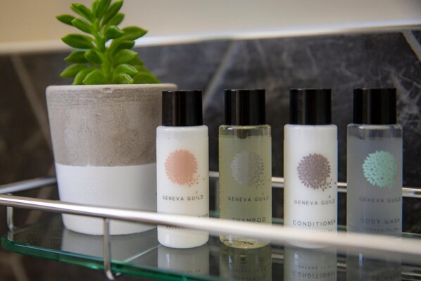 A bathroom shelf with four bottles of Geneva Guild products: shampoo, conditioner, body wash, and lotion. - The Princess Victoria (London)
