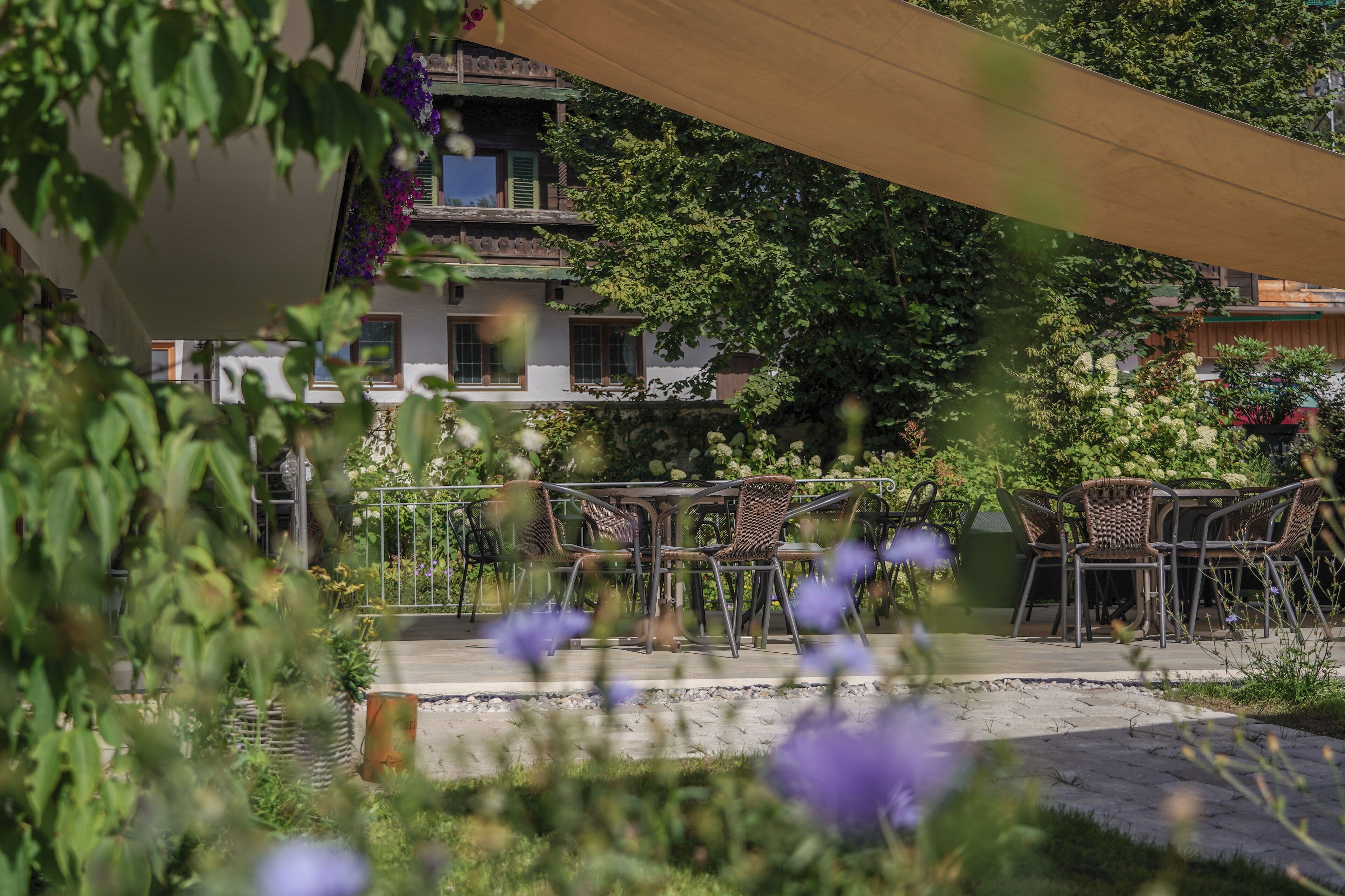 Eine sonnige Terrassenanlage mit Korbstühlen und einem Tisch, umgeben von üppiger Bepflanzung und Blumen.