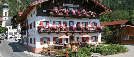 En traditionell alpbysbyggnad med en balkong som är prydd med blommor, en skylt som visar att det är en restaurang, och en utsikt över ett berg i bakgrunden.