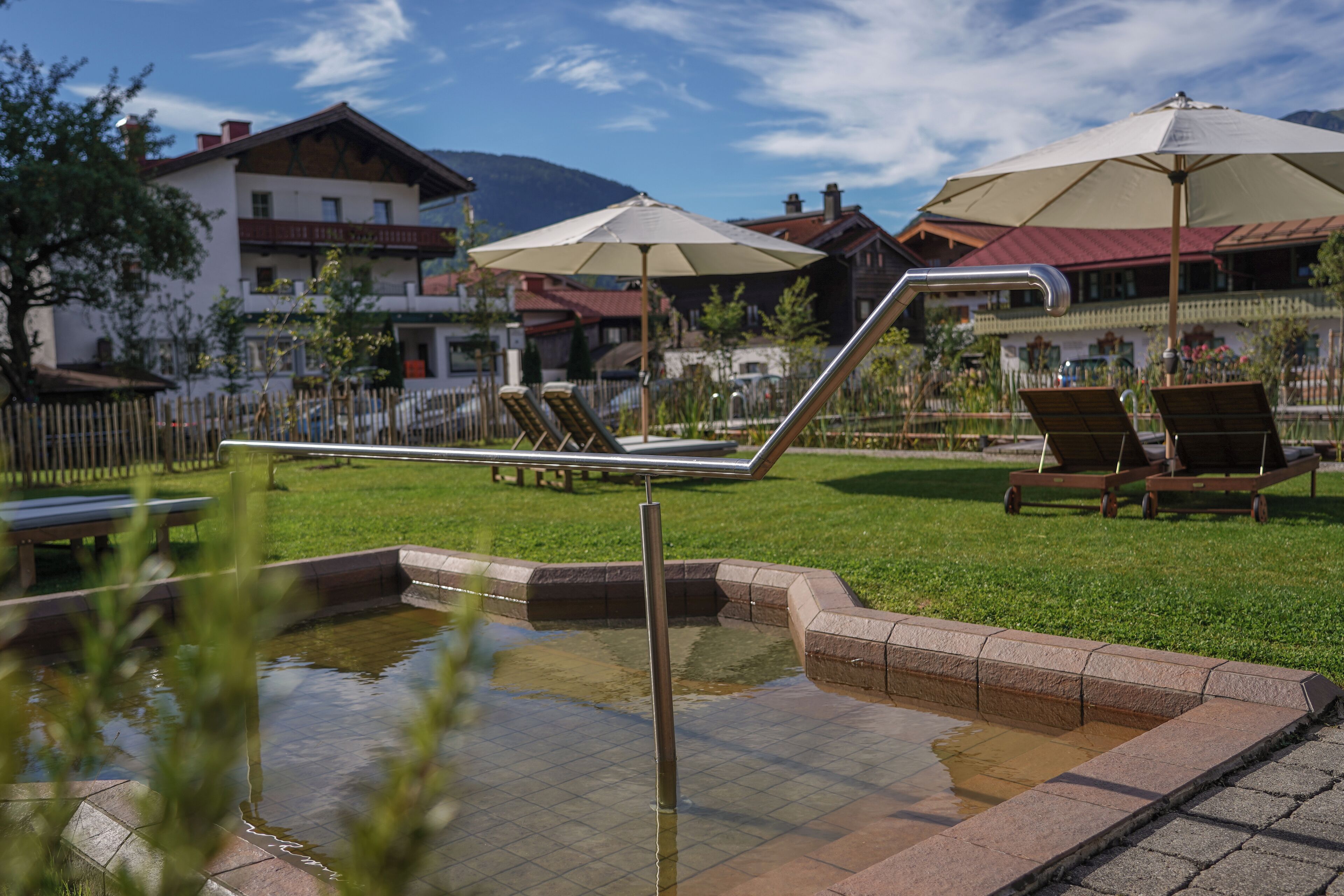 A pool area with lounge chairs and umbrellas, surrounded by a fence and buildings in the background.