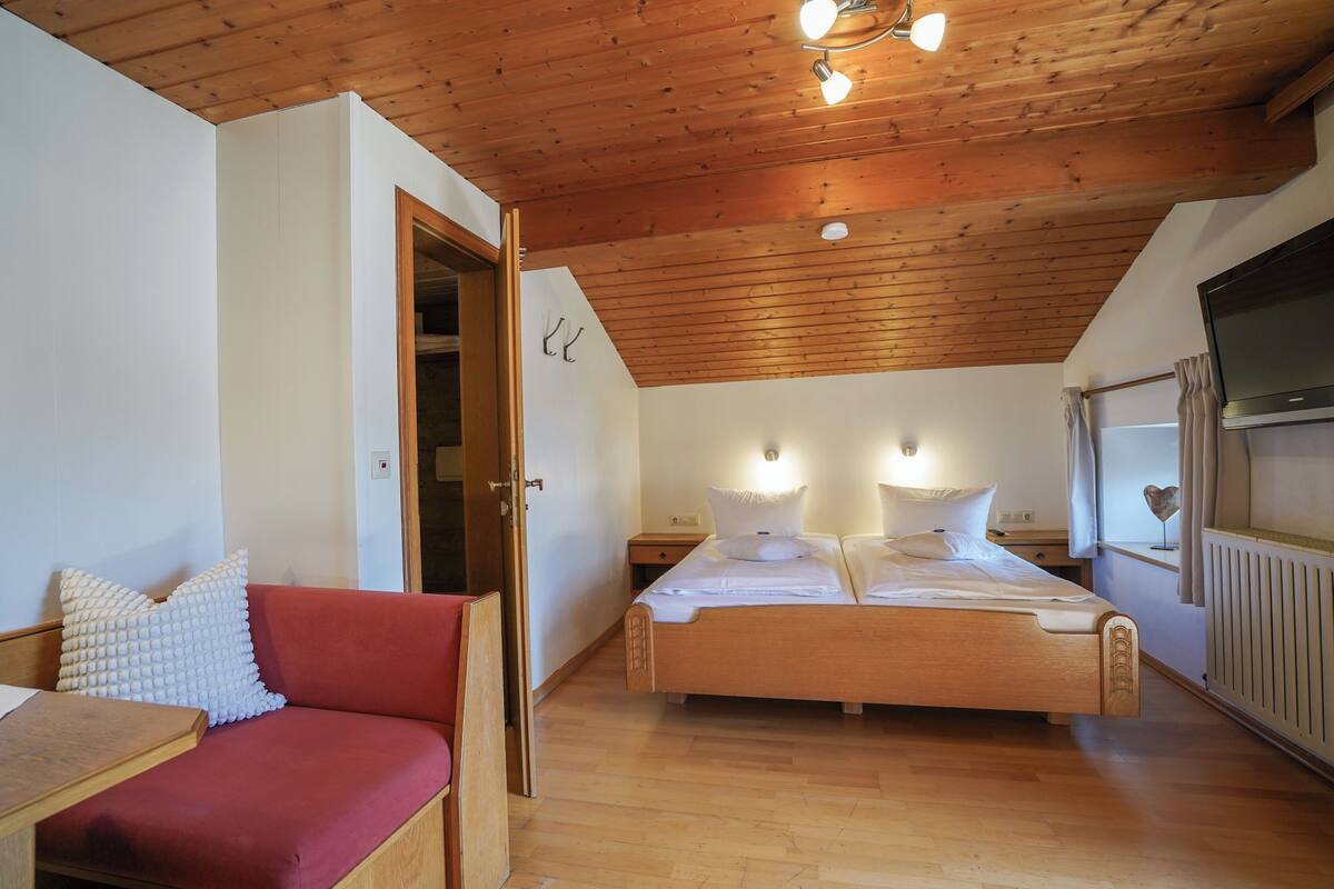 A bedroom with a wooden ceiling, a bed with white bedding, a red sofa, and a television on the wall.