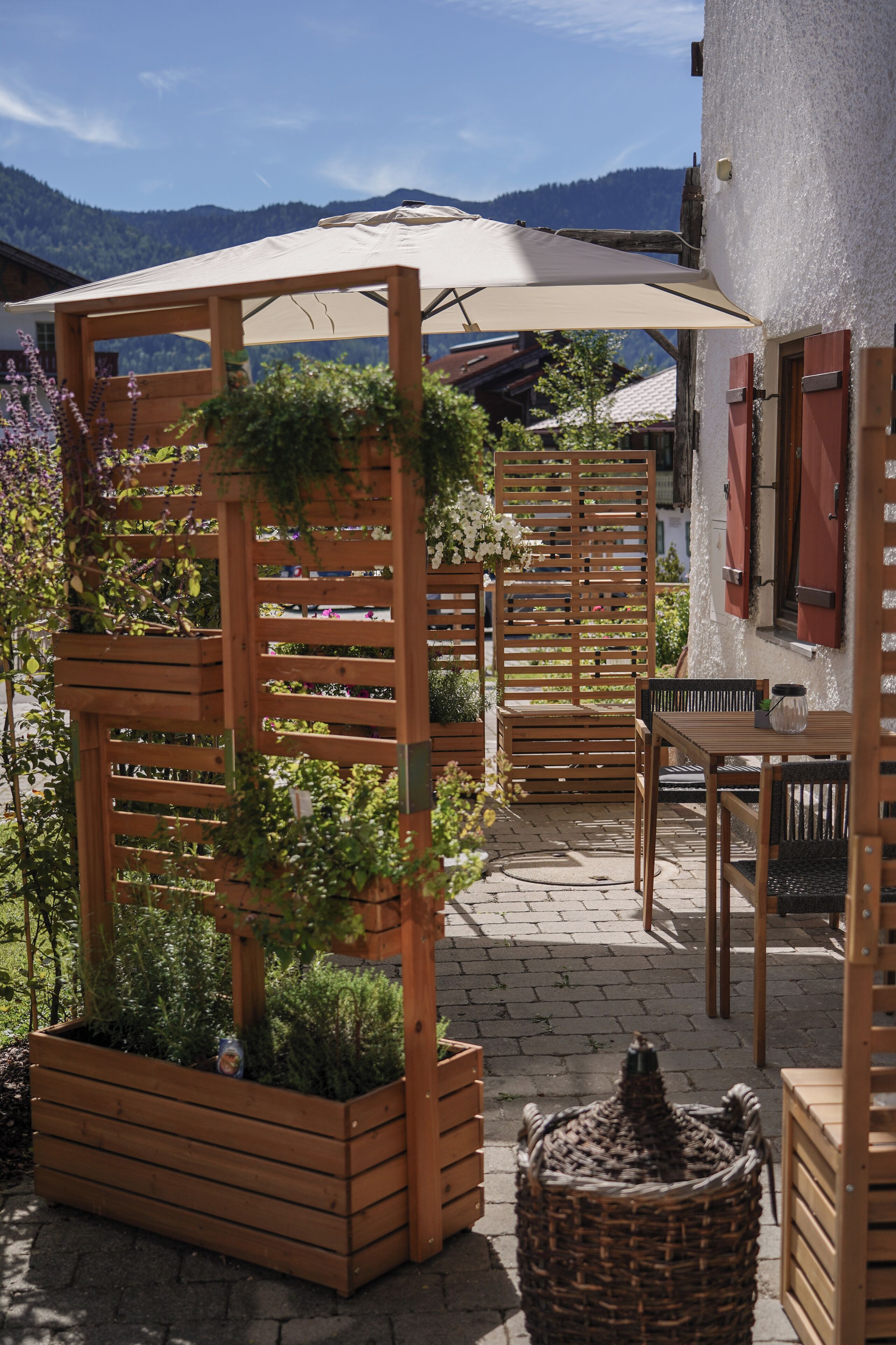 A wooden pergola with climbing plants, a table with chairs, and a wicker basket on a paved patio.