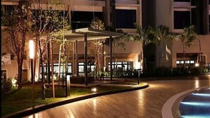 Vista nocturna de un vestíbulo de hotel con una zona de estar techada, una piscina y un camino bien iluminado.