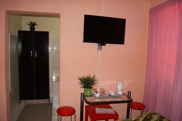 A small room with a TV mounted on the wall, a table with a plant, a coffee maker, and two red chairs. - RIO GUEST HOUSE LS (Maseru)