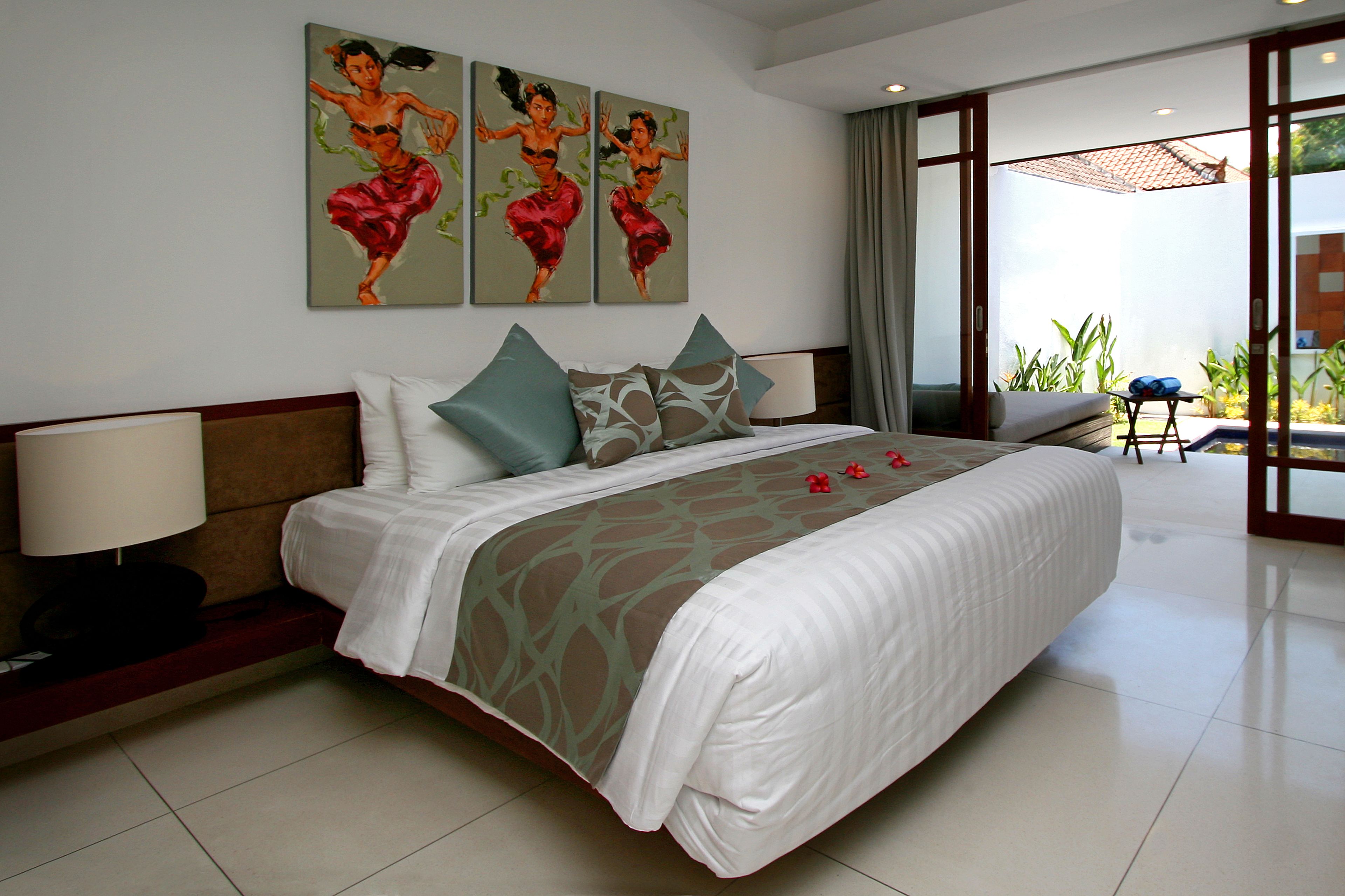 A hotel room with a large bed, a nightstand with a lamp, and three paintings on the wall.