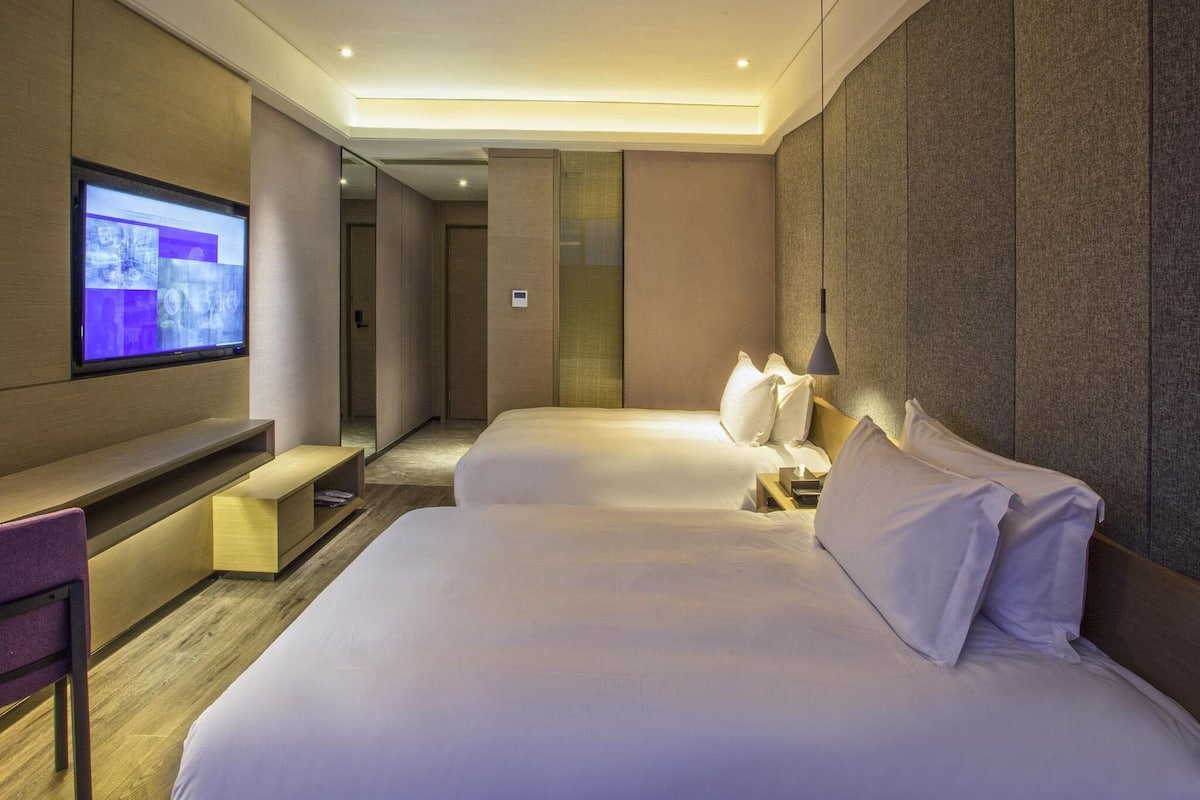 A hotel room with a TV, two beds, a desk, and a bench.