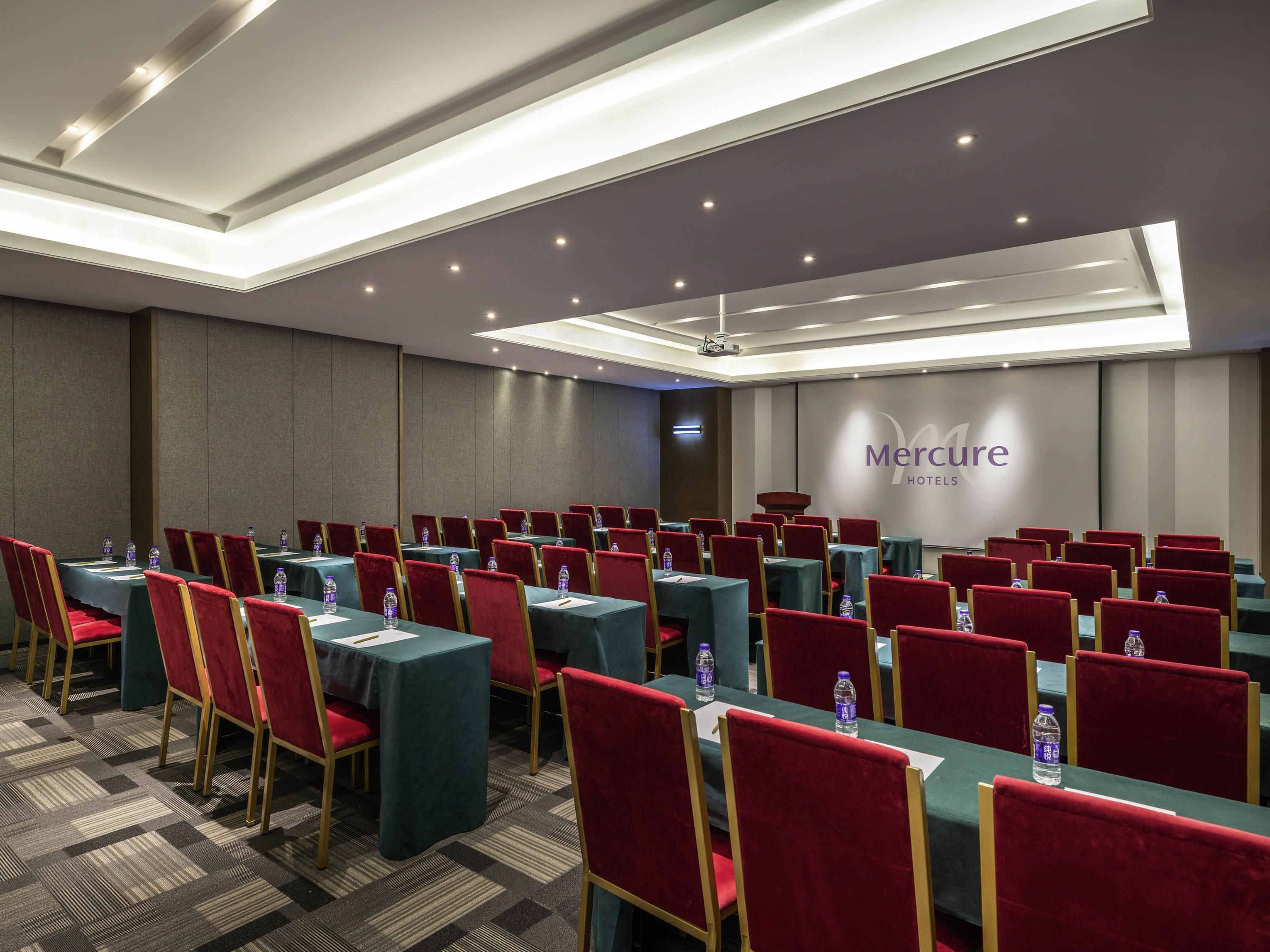 A conference room with rows of tables and chairs, a ceiling with recessed lighting, and a wall with the hotel name.