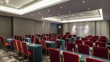 A conference room with rows of tables and chairs, a ceiling with recessed lighting, and a wall with the hotel name.
