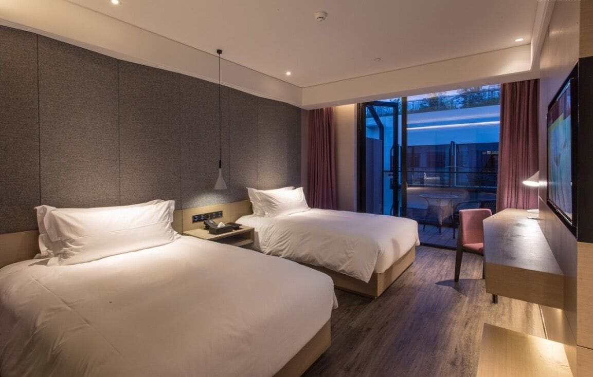 A modern hotel room with two beds, a desk, and a view of the outdoors.