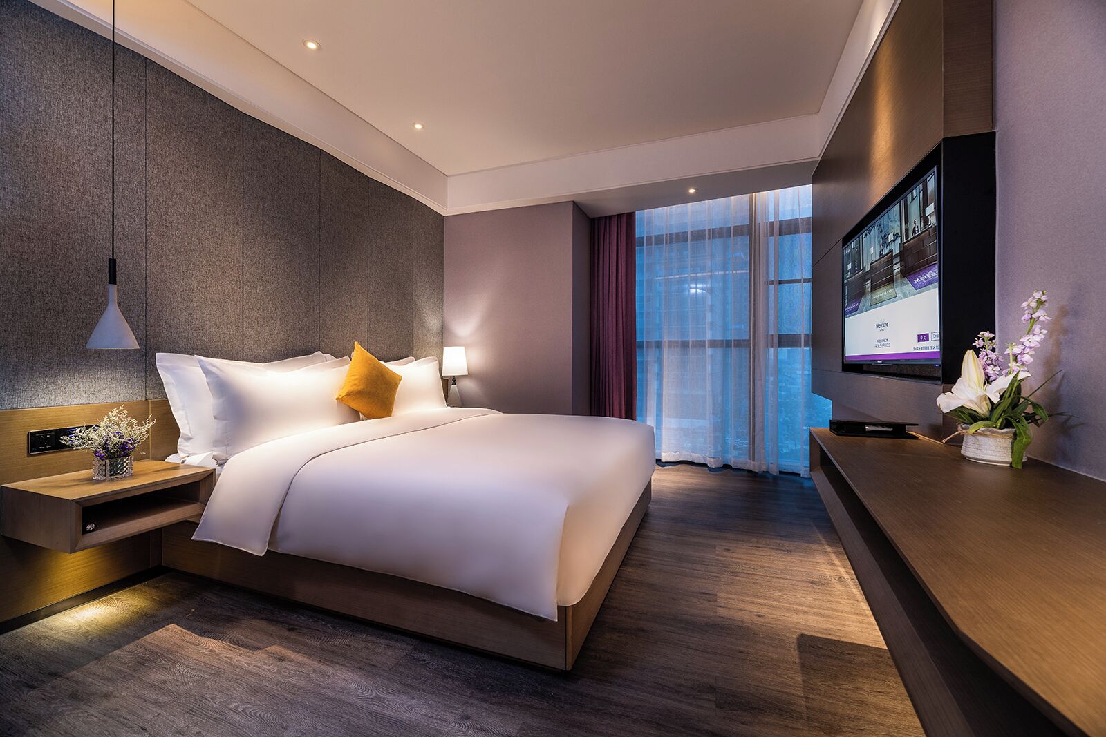 A modern hotel room with a large bed, a TV, a desk, and a view of the city.