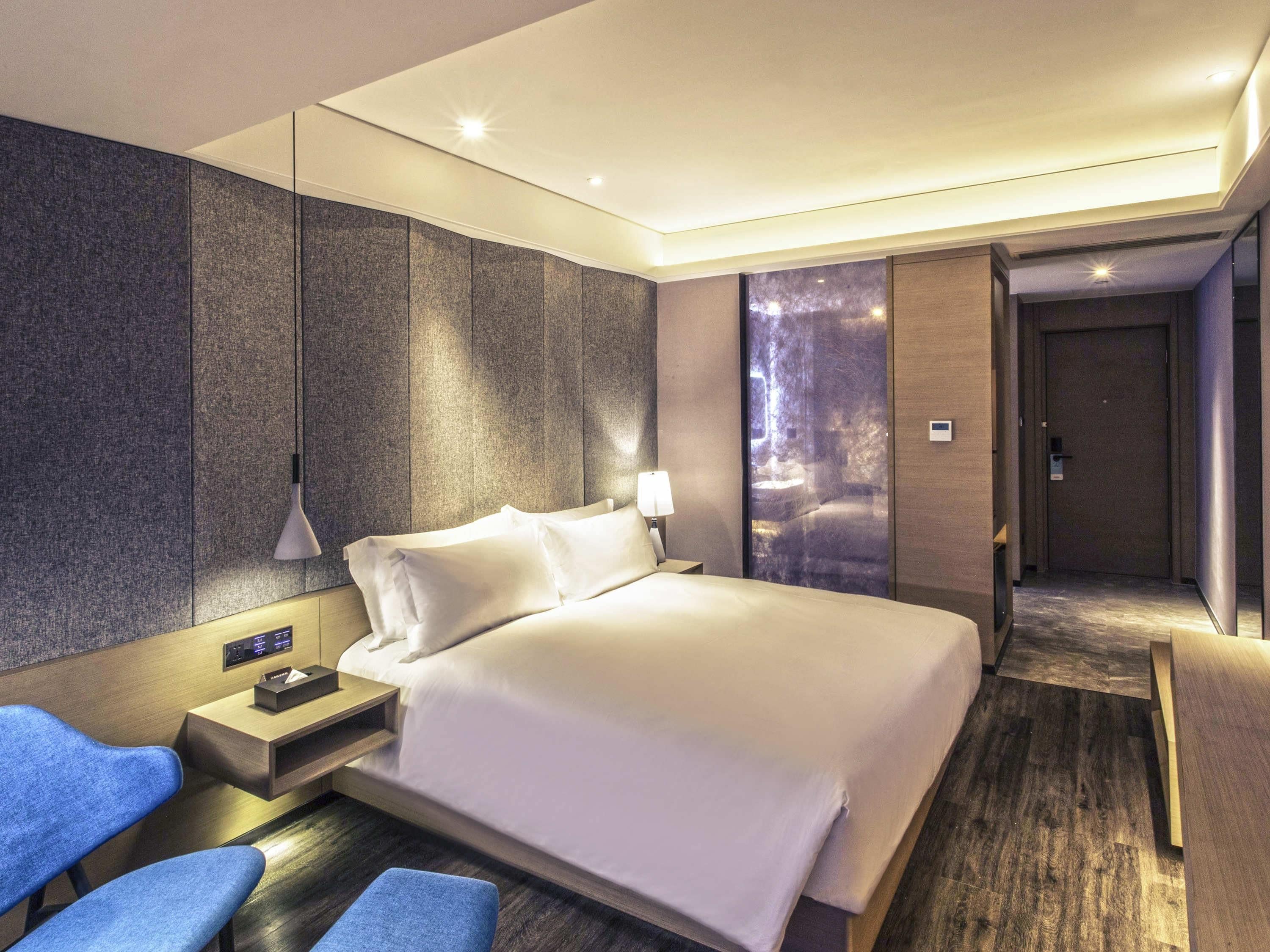 A hotel room with a bed, bedside table, lamp, and a view of a stone wall.