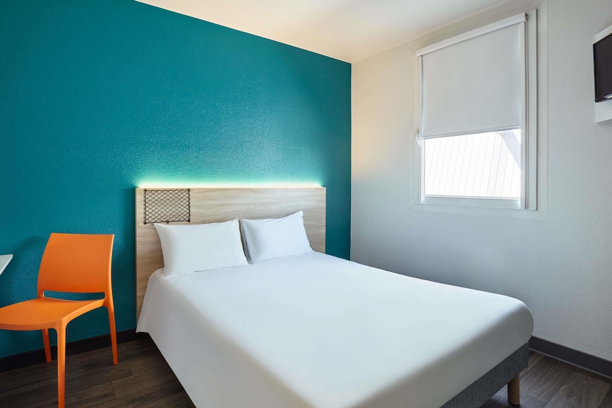 A hotel room with a bed, a chair, a window, and teal walls.