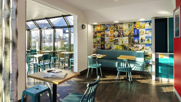 A cozy cafe with wooden tables and teal chairs, a green sofa, and a wall decorated with colorful posters.