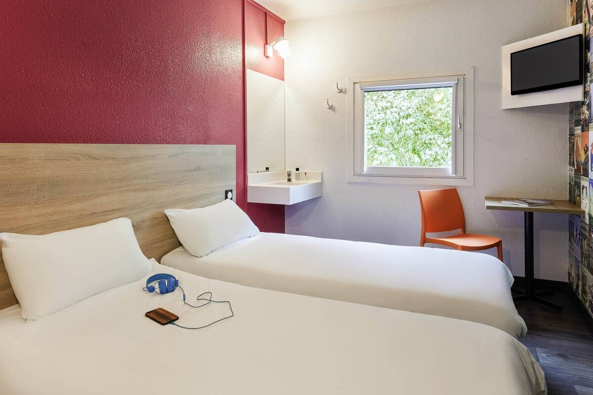 A hotel room with two beds, a desk, a chair, a TV, and a window with a view of greenery.