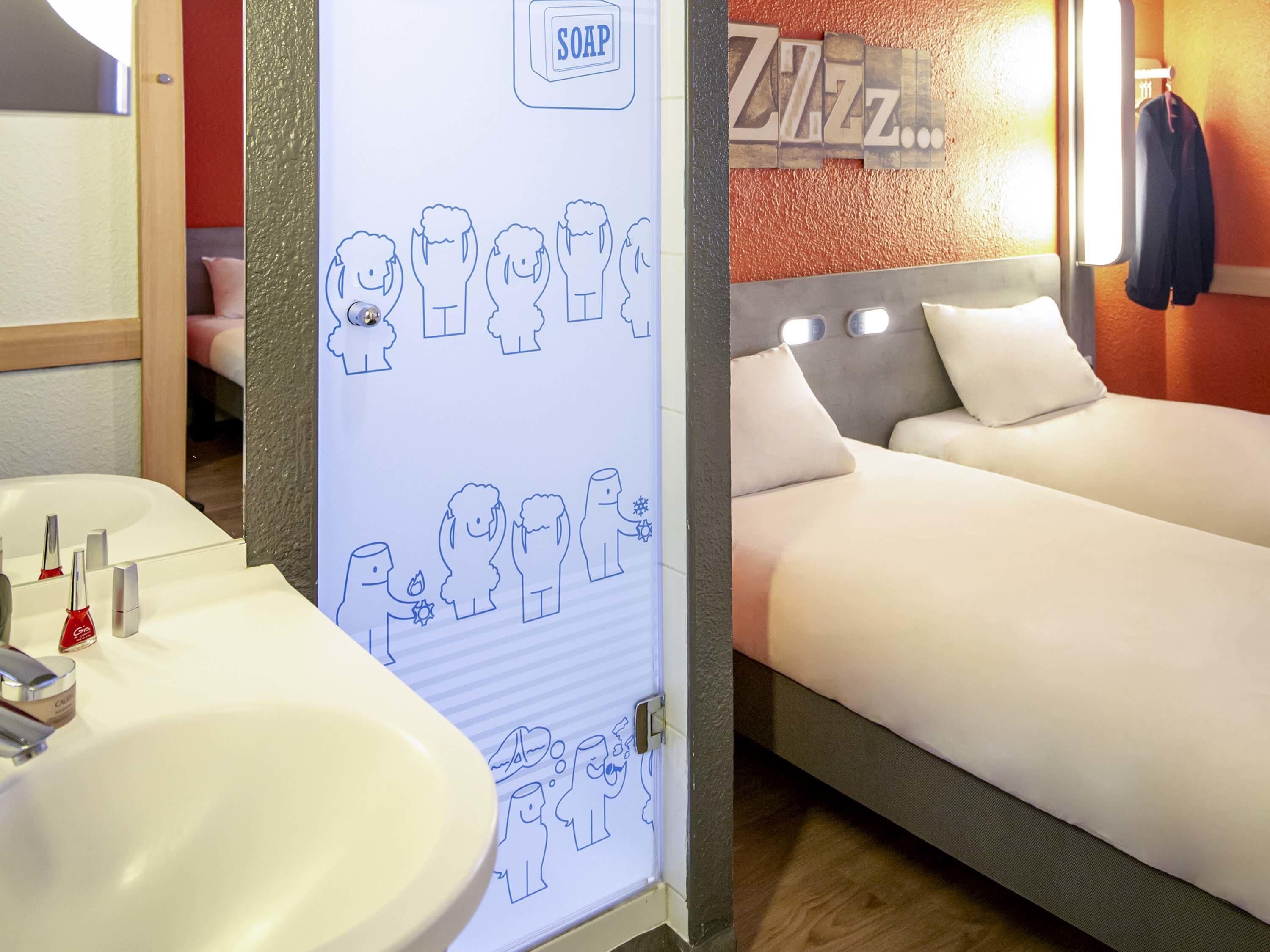A hotel room with a bed, a bathroom with a sink and toiletries, and a wall with a soap dispenser and a sign.