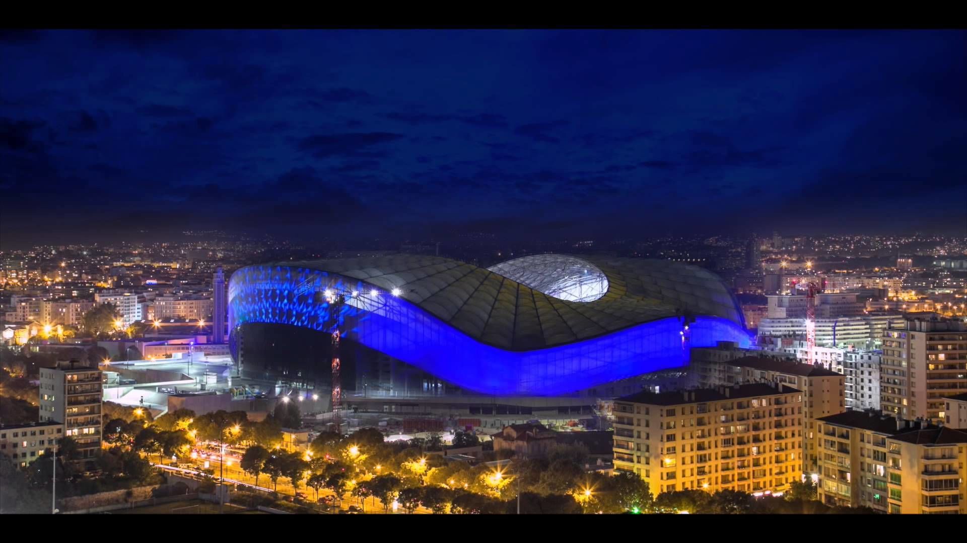 A modern stadium with a distinctive curved roof, surrounded by urban buildings and illuminated streets.