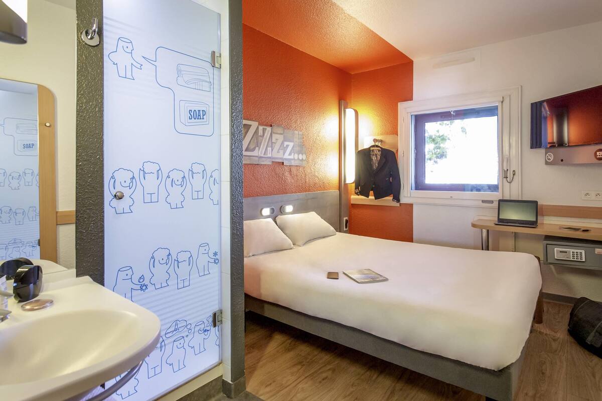 A hotel room with a bed, a bedside table with a laptop, a mirror, a window, and a wall with a soap dispenser.