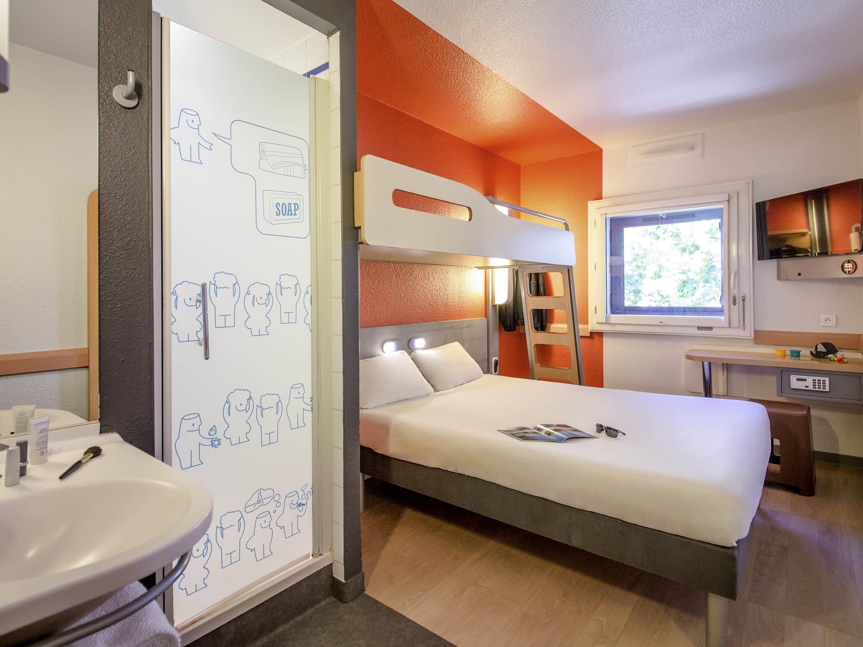 A hotel room with a bunk bed, a bedside table, a window with a view, a sink, and a mirror.